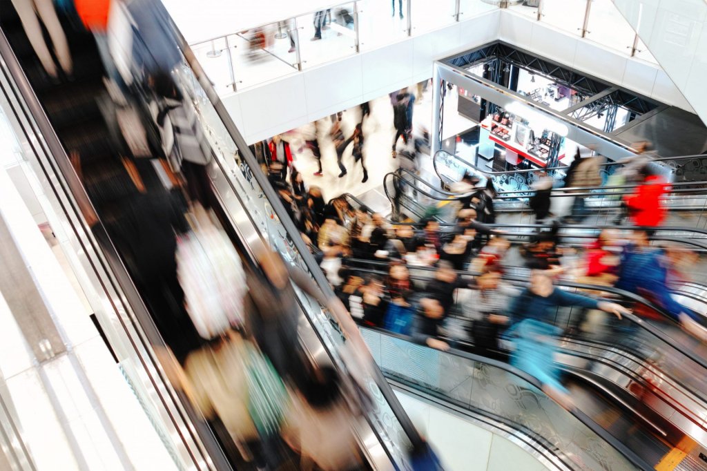 Long exposure photograph of shoppers on escalators in a busy shopping centre.
